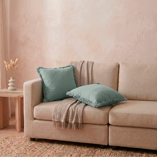 Combinación relajante de cojines decorativos en el sofá, destacando la delicada funda azul claro de algodón natural con flecos de Oma Home.